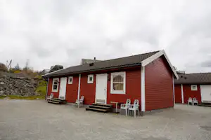 Rotes Ferienhaus mit Terrasse und Stühlen vor dem Eingang, Kiesweg und weitere Gebäude auf dem Campingplatz.