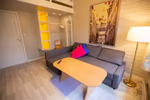 Graues Sofa mit bunten Kissen und Holztisch in einem hellen Wohnbereich, daneben Schrank und Wandbild.