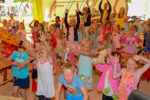 Kindergruppe vor einem gelben Zelt auf dem Campingplatz, mit Tischen und Stühlen im Hintergrund.