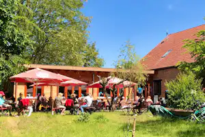 Campinghof mit rotem Sonnenschirm, Sitzgruppen und Stühlen vor einem Gebäude, umgeben von Rasen und Bäumen.