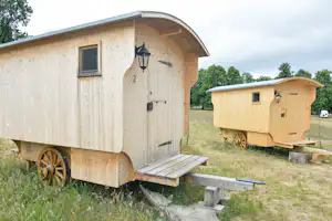 Zwei rustikale Wohnwagen auf einer Campingwiese, mit Holztreppe, Bank und Wagenrädern vor Bäumen und Wiese.