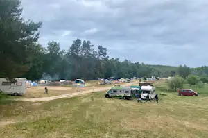 Campingplatz mit Wohnwagen und Zelten auf einer Wiese, Schotterweg und geparkten Autos, im Hintergrund Bäume und Wald.