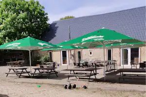 Grüne Sonnenschirme und Gartenmöbel auf einer Terrasse vor einem Backsteingebäude, im Hintergrund ein Ziegeldach und Bäume.