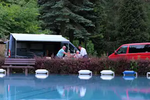 Mobilhome am Pool mit roten und schwarzen Autos, Holzstufen und Büschen im Hintergrund, Bäume im Garten.