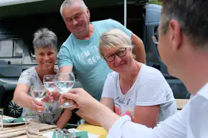 Camping-Tisch mit Gläsern, vier Personen sitzen vor Wohnwagen und unterhalten sich.