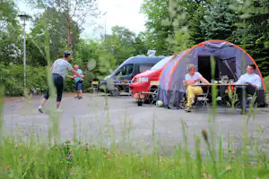 Campingzelt mit Sitzgruppe und Tisch auf dem Stellplatz, im Hintergrund rote Autos und Bäume im Grünen.