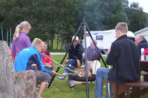 Grillabend am Campingplatz: Personen sitzen am Feuerkorb, im Hintergrund Wohnwagen und Bäume im Grünen.