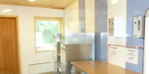 Küchenzeile im Campinggebäude mit Edelstahlspüle, Schränken, Holzdecke und Fenstern zur Umgebung.