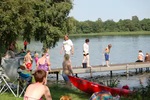 Familie am Steg am See: Kinder und Erwachsene am Holzsteg, Kajaks im Vordergrund, Bäume am Ufer.