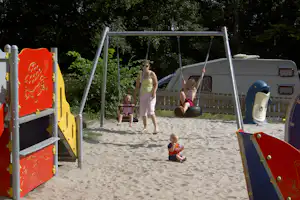 Spielplatz mit Schaukeln und Sandboden, im Hintergrund Wohnmobil und Holzzaun, umgeben von Bäumen.
