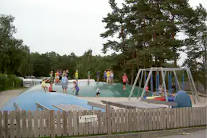 Kinder auf einem Spielplatz mit großer grüner Rutsche, Sprungbereich und Klettergerüst, im Hintergrund Bäume und Wohnwagen.