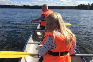 Kinder im Boot mit Schwimmwesten auf dem See, gelbe Paddel und Ruder, im Hintergrund Wald und Uferlinie.