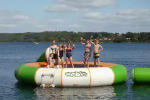 Aufblasbares Badeboot auf einem See, Kinder im Schwimmanzug winken, im Hintergrund bewaldete Hügel und Uferhäuser.