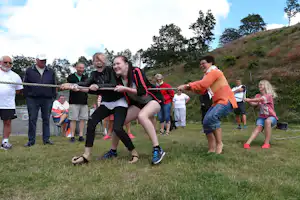 Campinggruppe beim Seilziehen auf einer Wiese, mehrere Personen halten das Seil, im Hintergrund Bäume und ein Hang.