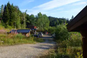 Campingplatz mit Kiesweg, Wohnwagen und Autos, umgeben von Bäumen und Sträuchern, im Hintergrund ein Gebäude.
