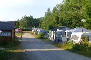 Campingplatz-Gasse mit Wohnwagen und Zelten, Laternen und Bäumen am Rand, Gebäude im Hintergrund.
