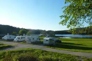 Stellplätze am See mit Wohnmobilen und Vorzelten, Kiesweg und Rasenfläche, im Hintergrund Wald und ruhiges Wasser.