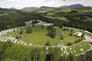 Luftaufnahme eines Campingplatzes mit vielen Stellplätzen, Wohnmobilen und Zelten, umgeben von Wiesen, Bäumen und Hügeln im Tal.