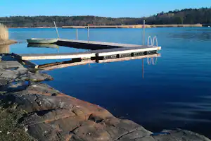 Bootssteg am See mit festgemachtem Boot, Holzplanken und Metallgeländern, im Hintergrund Wald und Uferlinie.