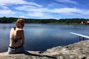 Frau sitzt am Seeufer auf Felsen, Blick auf ruhiges Wasser, im Hintergrund Wald und ein rotes Gebäude.