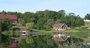 Rotes Ferienhaus am See mit Spiegelung im Wasser, umgeben von Wiesen, Bäumen und einem weiteren Gebäude am Ufer.