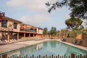 Schwimmbad mit Poolrand, Liegen und Stühlen, dahinter ein mehrstöckiges Gebäude mit Balkon und Holzgeländer.