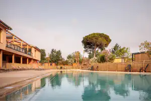 Schwimmbecken eines Campingplatzes mit Holzterrasse am Gebäude, umgeben von Bäumen und Zäunen.