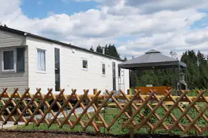 Weiße Wohnwagenanlage mit Holzlattenzaun und Rasen, im Hintergrund ein weiteres Gebäude und Bäume.