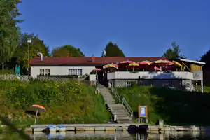 Campinghaus mit rotem Dach, Terrasse und Treppe zum Ufer, am See mit Spiegelung im Wasser und grünen Büschen.