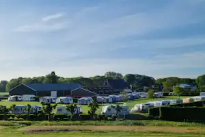 Campinganlage mit vielen Wohnwagen auf grünen Stellplätzen, Hecken und Bäumen im Hintergrund, große Hallen und Gebäude.