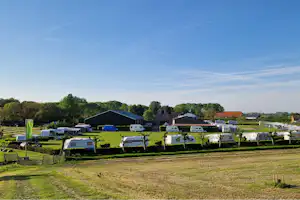 Reihe von Wohnwagen auf einem grünen Campingplatz, mit Hecken, Bäumen und großen Hallen im Hintergrund.