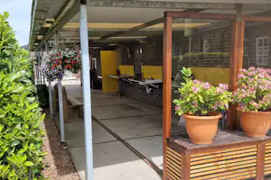 Grillplatz unter einem Holz-Überdachung mit gemauerten Wänden, gelben Spülbecken und Blumenbeeten im Vordergrund.