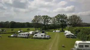 Wohnmobile und Autos auf einer Campingwiese, dazwischen Zelte, Bäume und ein Weg unter bewölktem Himmel.