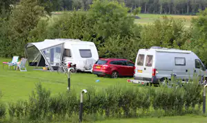 Wohnwagen mit Vorzelt, daneben rotes Auto und weißes Wohnmobil auf einem Campingplatz auf grünem Rasen, umgeben von Bäumen.