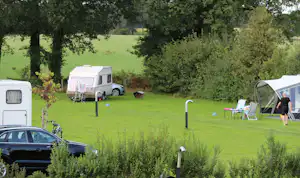 Wohnwagen auf einem grünen Campingplatz, daneben Autos und Campingstühle, im Hintergrund Bäume und Wiese.