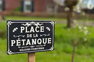 Schild „Place de la Pétanque“ auf einem Holzpfosten, im Hintergrund Rasen und Gebäude am Campingplatz.