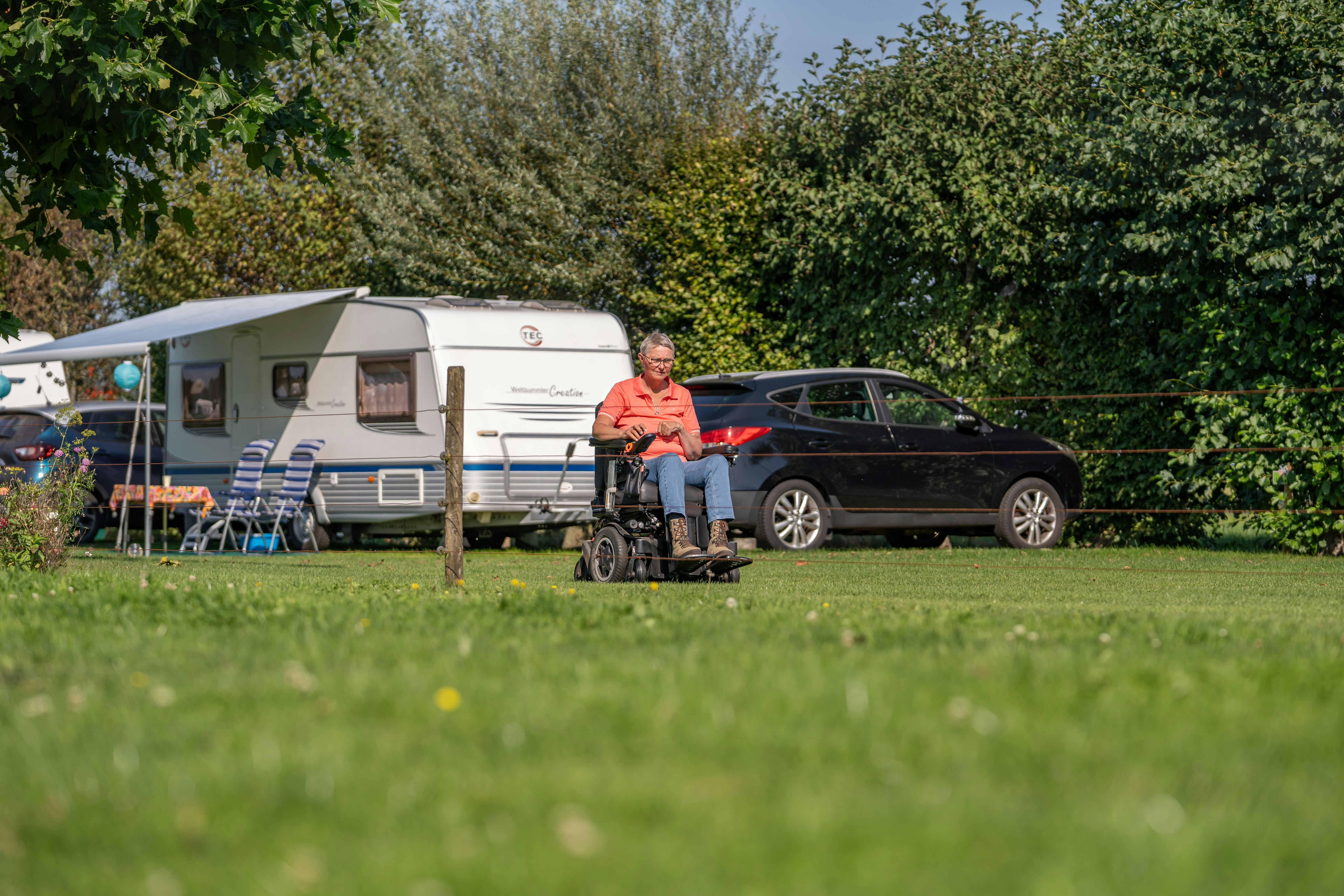 Wohnwagen und Auto auf dem Campingplatz, daneben Rollstuhl und Sonnenschirm, im Hintergrund Hecken und Rasen.