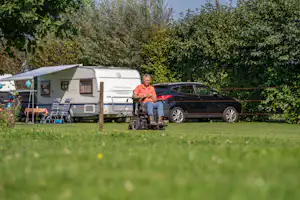 Wohnwagen und Auto auf dem Campingplatz, daneben Rollstuhl und Sonnenschirm, im Hintergrund Hecken und Rasen.
