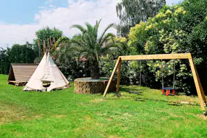 Tipi-Zelt auf einer grünen Wiese, Holzschaukel und Steinmauer im Garten mit Palmen und Büschen.