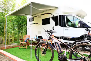 Wohnmobil mit Markise, zwei Fahrräder vor dem Fahrzeug und Holzstühlen am Stellplatz im Grünen.