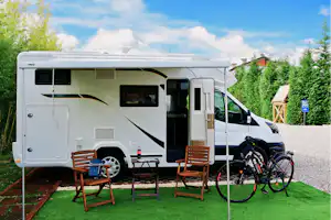 Wohnmobil mit Markise, zwei Holzstühlen und Tisch am Stellplatz, Fahrrad am Heck, gepflegter Rasen und Hecken im Hintergrund.