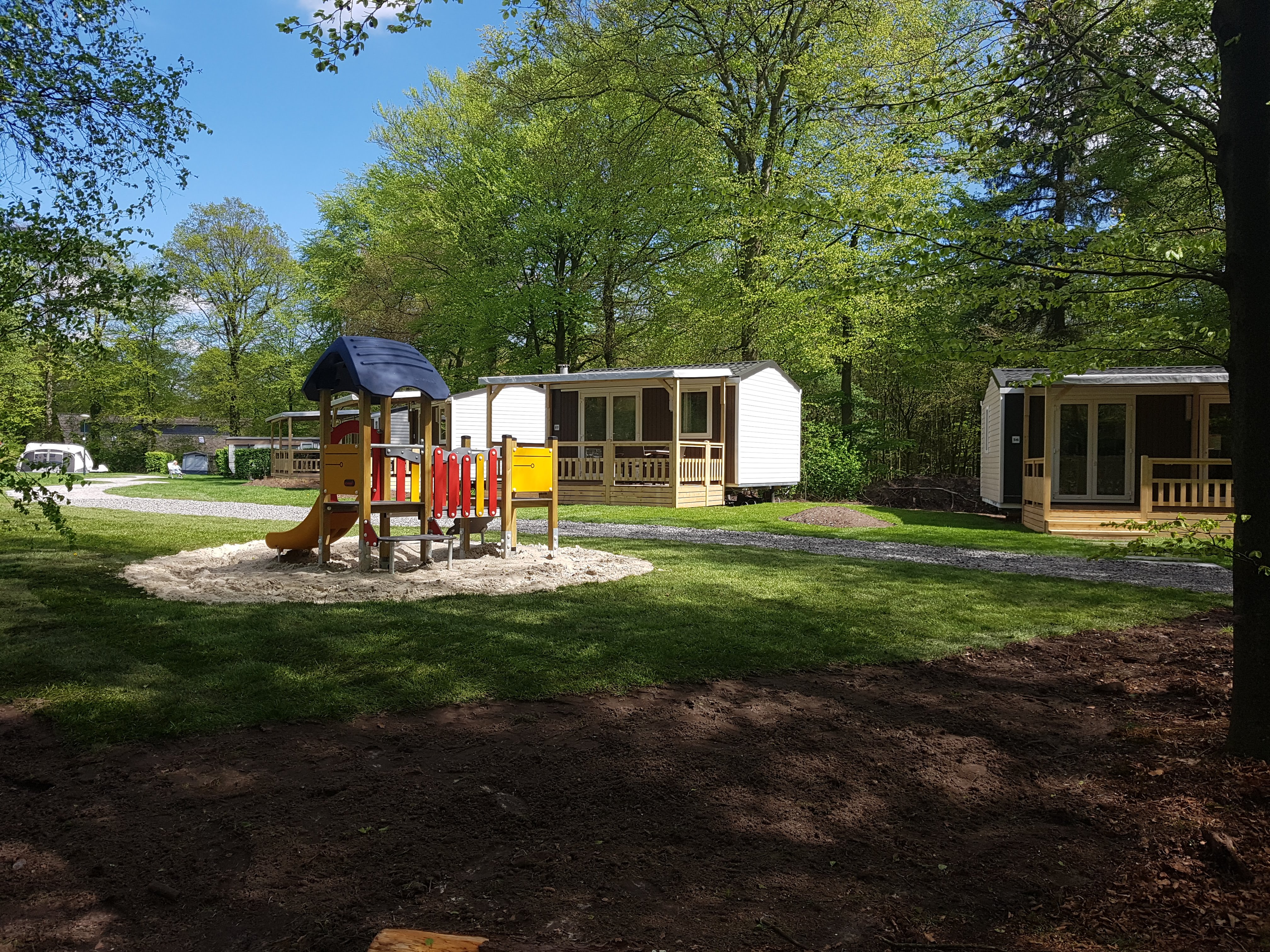 Holzbungalow mit Terrasse und Spielgeräten auf einem Campingplatz, umgeben von Bäumen und Rasenflächen.