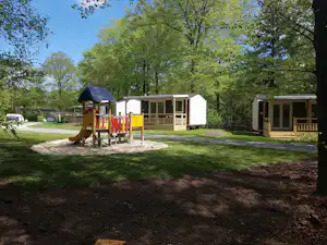 Holzbungalow mit Terrasse und Spielgeräten auf einem Campingplatz, umgeben von Bäumen und Rasenflächen.