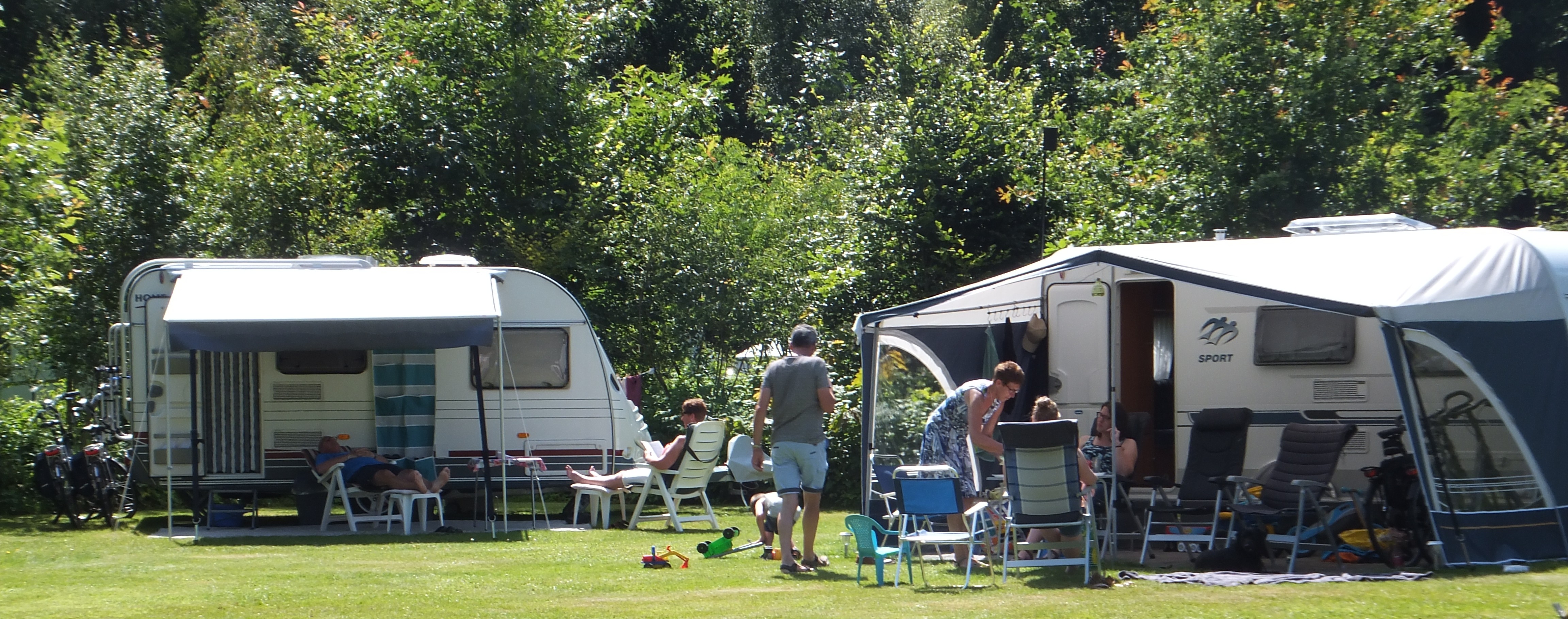 Zwei Wohnwagen auf einem Campingplatz mit Rasen, Sonnenschirmen und Stühlen vor den Eingängen, umgeben von Bäumen.