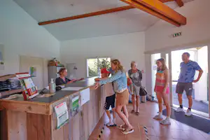 Rezeption im Campinghaus mit mehreren Urlaubern, Holztheke und Infomaterial an der Wand, große Fenster im hellen Raum.