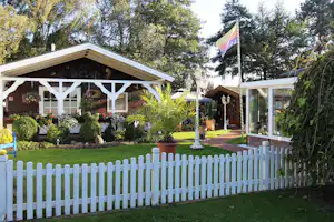 Mobilheim und Wohnwagen auf dem Campingplatz mit weißem Zaun, Gartenpflanzen und Terrasse im Grünen.