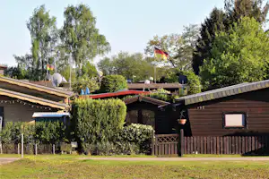 Holzbungalow auf einem Campingplatz mit Rasen, Hecken, Sitzbereich und Flagge im Garten.