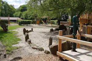Sandspielbereich mit Klettergerüst, Holzpfosten und Rutsche, im Hintergrund Hecken und Bäume auf dem Campingplatz.