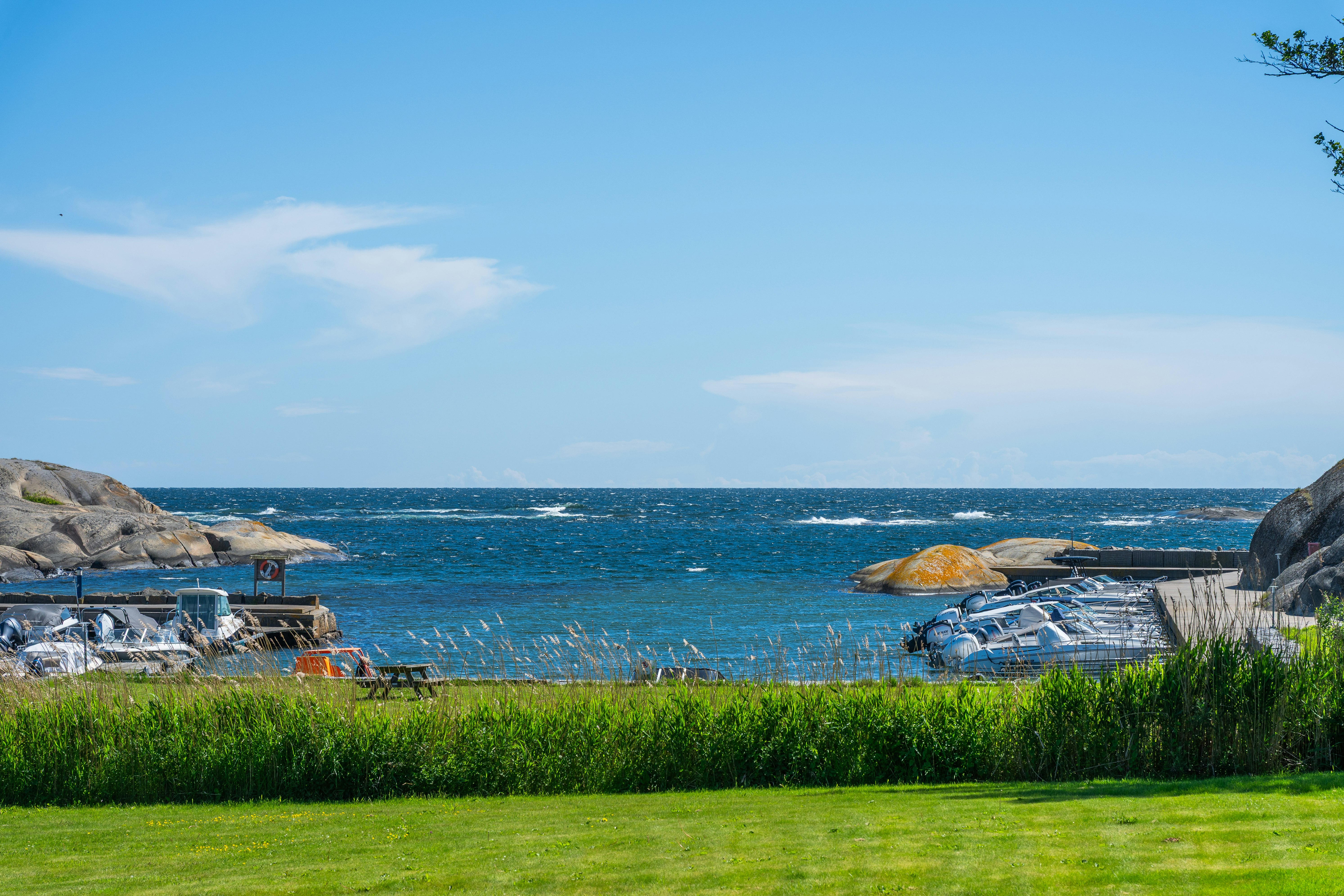 Blick auf das Meer mit Strandliegen und Felsen am Ufer, davor grünes Gras und ein Campingplatzbereich.