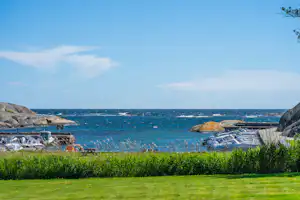 Blick auf das Meer mit Strandliegen und Felsen am Ufer, davor grünes Gras und ein Campingplatzbereich.